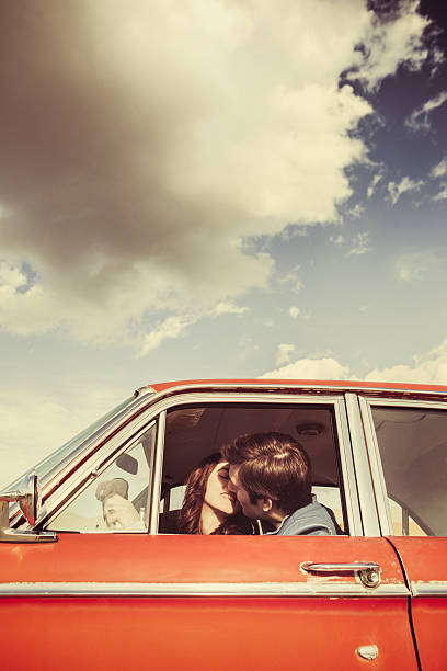 A young man and woman are visible kissing through the driver window of their classic 1960s car.  Vintage styling and colors.  Wide and low angle to emphasis the blue sky and clouds.  Vertical with copy space.