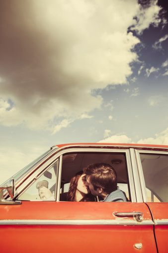 A young man and woman are visible kissing through the driver window of their classic 1960s car.  Vintage styling and colors.  Wide and low angle to emphasis the blue sky and clouds.  Vertical with copy space.