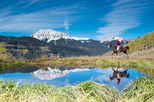 man horseback riding is reflected in the water of a pond with snowcapped rugged mountain peaks in the background.  such beautiful nature scenery and outdoor sports and recreation can be found in the san juan range of the colorado rocky mountains, durango.  horizontal composition.