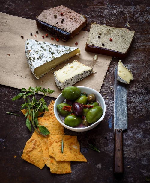 Front view of various types of cheese like Emmental cheese, cottage cheese, manchego cheese, parmesan cheese, brie cheese, cured and semi-cured cheeses on a bluish tint background. The composition includes black and green olives, an olive oil bottle, rosemary, a honey jar with a honey dipper, a pepper shaker and pepper. Predominant colors are blue and yellow. Low key DSLR photo taken with Canon EOS 6D Mark II and Canon EF 24-105 mm f/4L