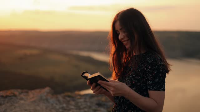 A girl in a dress reads a book in the sun. A young woman reads the Bible in the open air, holding the Bible in her hands and studying the word of God at sunset on the top of the mountain.