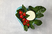 Piece of curd goat cheese and red cherry tomatoes on branch with green spinach leaves in heart shaped blue bowl on gray concrete table. Top view with copy space