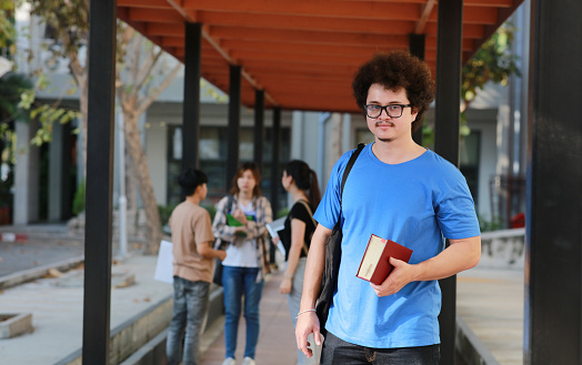 Banner size photo of diverse teenagers or young adults walking outdoors on school campus. Hispanic, Caucasian, and African American students are smiling while walking together to class. They are wearing trendy casual clothing and backpacks, and are carrying school books.
