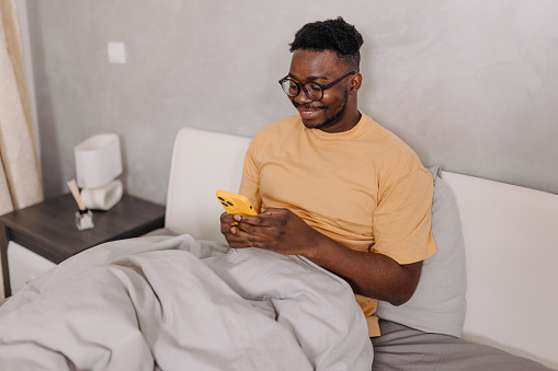 Exhibiting a strong sense of connectivity, this young man wakes up and instinctively reaches for his phone, ensuring he stays connected and informed right from the start, even before stepping out of bed