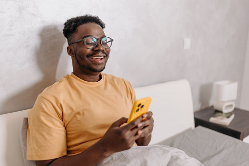 Embracing tech-savvy mornings, this young African American man utilizes his phone as a tool to kickstart his day, engaging in tasks such as checking emails, planning his schedule, or pursuing personal interests, all while enjoying the cozy comfort of his bed