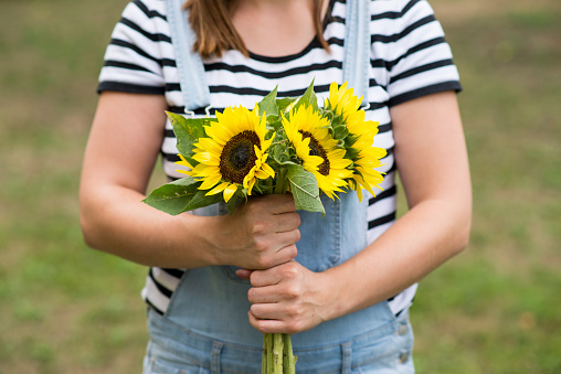 Anonymous young woman in jean overalls and a striped shirt is holding a beautiful and bright bouquet of sunflowers from the farmers market.