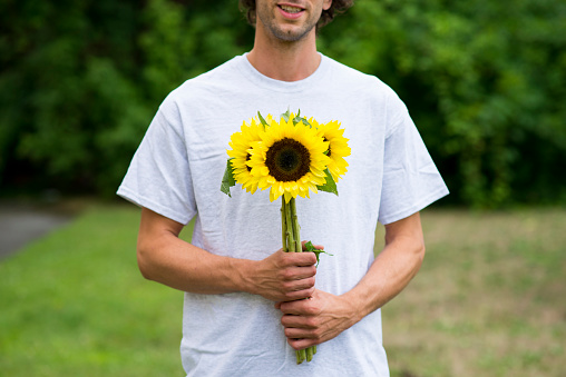 Anonymous, smiling young man in a white shirt is holding a beautiful and bright bouquet of sunflowers from the farmers market outdoors to give to his date.