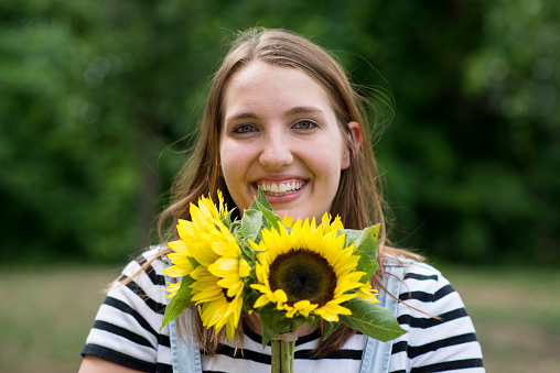 Smiling brunette young woman is holding up a beautiful bouquet of sunflowers in the summer outdoors.