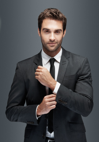 Gorgeous young man doing up his cufflinks and looking at you, isolated on grey - copyspace