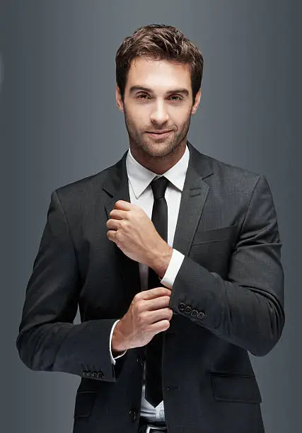 Gorgeous young man doing up his cufflinks and looking at you, isolated on grey - copyspace