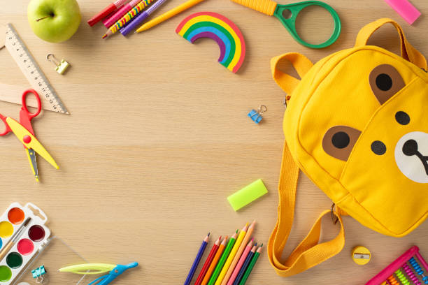 istock School materials scene. Top view of vibrant accessories, art kit, scissors, ruler, sharpener, magnifying glass, abacus, adorable bear-shaped bag, apples and more on wooden desk with blank area for ad 1554168062
