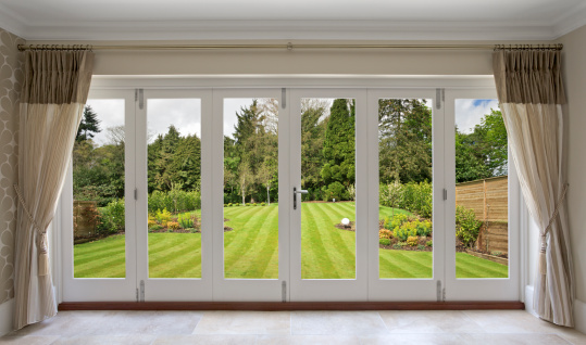 a set of concertina patio doors with silk curtains on either side and a view of a fabulous garden. A large nicely mowed lawn stretches away towards a backdrop of mixed trees.