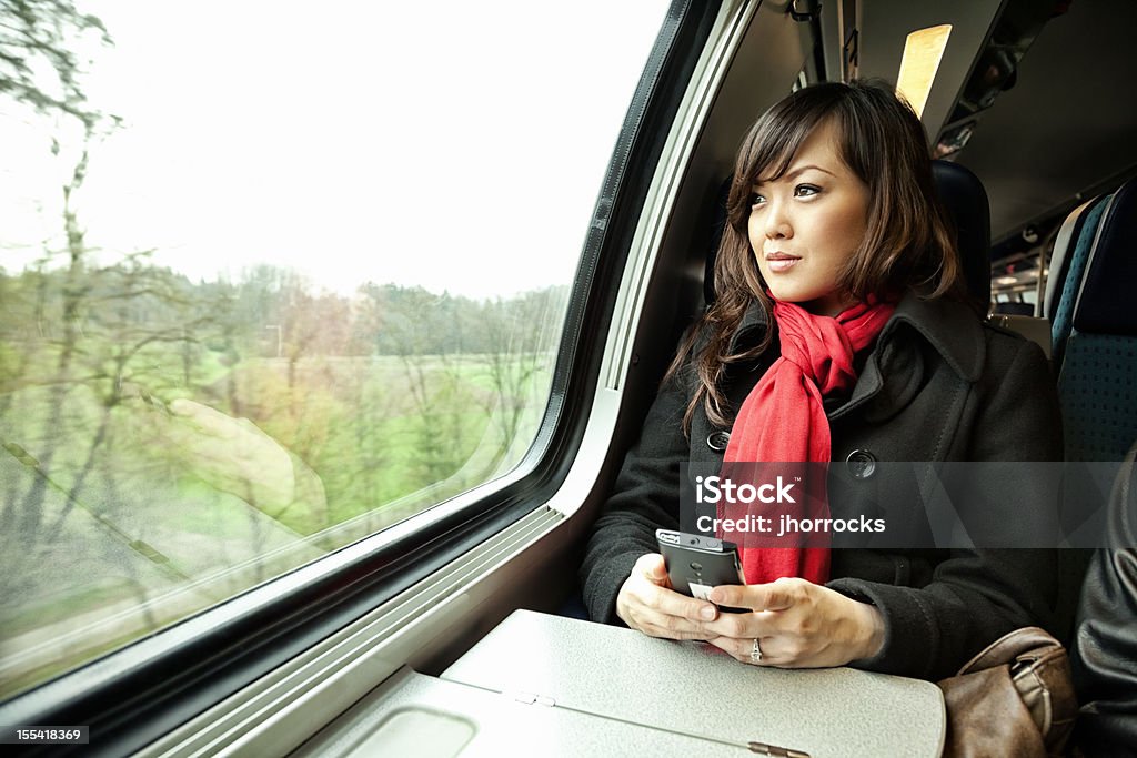 Young Asian Woman Traveling by Train Photo of an attractive young Asian woman holding her mobile phone as if waiting for a text message while riding a passenger train, looking out the window at the passing scenery. Mobile Phone Stock Photo
