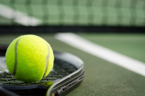 A black tennis racket and yellow tennis ball laying on the ground at a tennis court in early morning light.