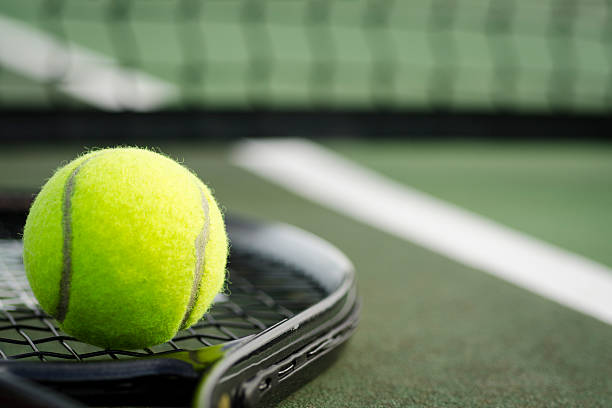 Tennis Ball and Racket on the Court Horizontal A black tennis racket and yellow tennis ball laying on the ground at a tennis court in early morning light. tennis ball stock pictures, royalty-free photos & images