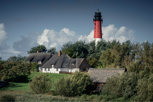 Pellworm, Germany - October 08, 2022: The Pellworm lighthouse behind thatched roof captain's houses under a blue sky. The lighthouse was completed in May 1907 after a year of construction. The height of the tower is 41.5 meters. It stands on a pile foundation consisting of 127 oak piles. The piles were driven into the ground to a depth of 14 meters to distribute the massive weight of 130 tons of cast steel.