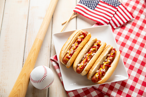 This is a photograph of three hot dogs on a white plate on a white wooden picnic bench. This is a great image for a Fourth of July picnic with a baseball and bat