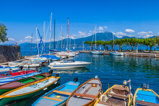 A row of colorful fishing boats and sailboats are lined in the small harbor of Torri del Benaco, a tiny village of extraordinary charm on the Veronese shore of Lake Garda. Tourists are visiting the historic town overlooking the beautiful lake, the largest lake of Italy.