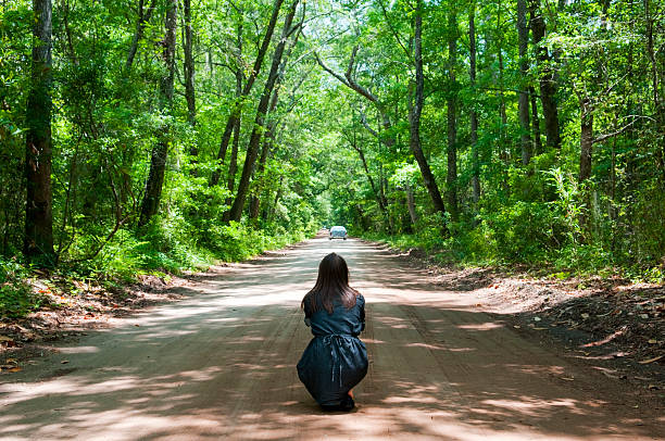 Young woman crouched and thinking on dirt road in summer A teenager or young woman crouched on a dirt road, looking at a car as it drives away sad girl crouching stock pictures, royalty-free photos & images