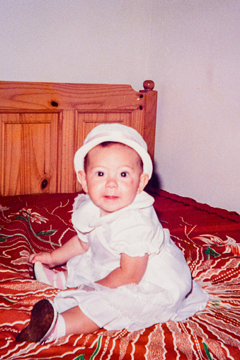 Vintage portrait of baby in white dress sitting on bed.