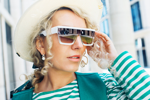 Portrait of an adult woman with blond curly hair wearing sunglasses and hat. City portrait close up.