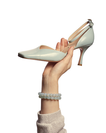 A female hand with round stone bracelets holding an elegant soft blue summer shoe with high heel isolated on a white background. Graceful hand showcasing footwear