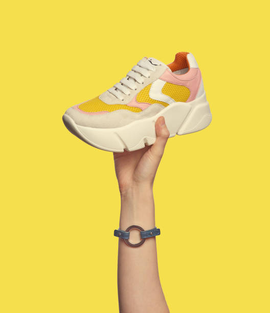 istock A female hand holding a trendy multicolor sneaker with a chunky platform isolated on a yellow background. Sport and active lifestyle. Stylish shoe presentation in hand 1554266733