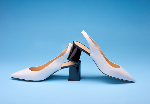 Elegant open-back white pumps with black shiny flare heels on a gradient blue background. One shoe stands on another. Copy space. Mock-up for design advertising for a shoe store