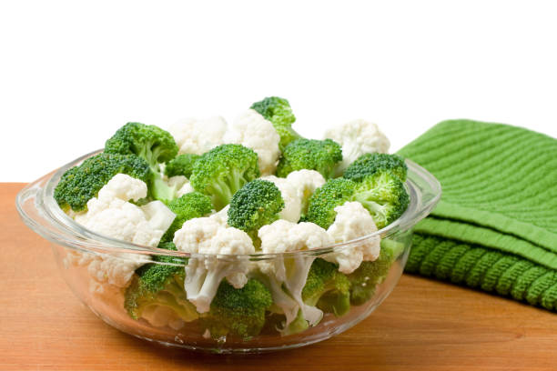 A glass bowl of cauliflower and broccoli on a wood cutting board.