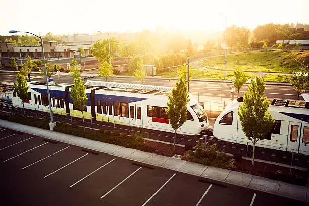 The MAX Line: Rail train public transportation in Portland Oregon traveling on the Clackamas route stops at a parking ride to pick up passengers.  Bright vibrant morning light with green and blue colors.  Horizontal with copy space.