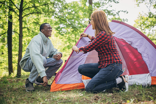 Multiracial couple, male and female hikers setting up a tent together in nature.