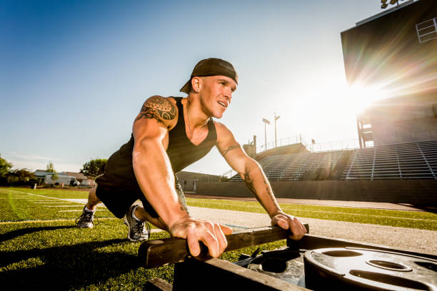Young man Pushing Weighted Sled in a gym style workout.  Man has a positive smile on his face, looking away from the camera in a sleeveless shirt and a ball cap on backwards.  He also has a large tattoo on his shoulder.