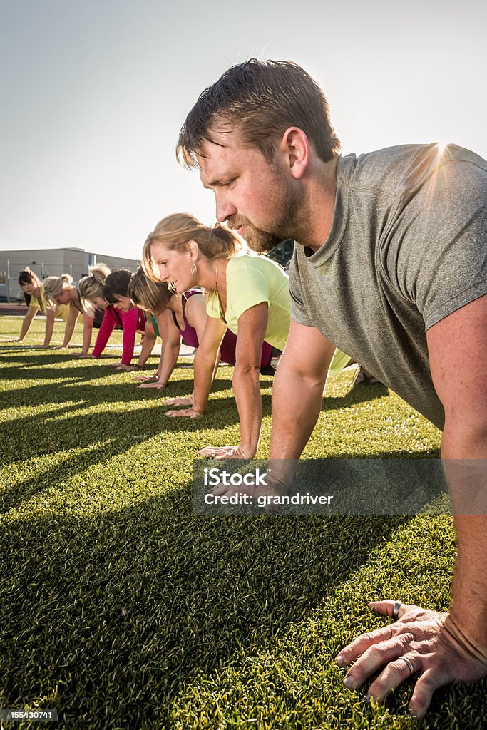 Line of people doing pushups or planks outside on grass Seven men and women wearing athletic clothing and sitting in the push-up position on a grassy field early in the day.  There are buildings and a blue and white sky behind them.  There is blue sky above them and long shadows on the freshly cut green grass. Outdoors Stock Photo