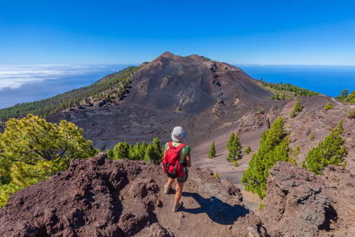 Woman admiring the beautiful volcanic landscape along the Volcano Route, maybe the most spectacular trek of La Palma that follows the ridge of the mountain from El Pilar to Fuencaliente, for a total of about 17 km. You can walk on the edge of a crater, on a land paved with stones and volcanic rocks, but also through the vegetation that gradually takes possession of the territory. And enjoy the panorama on both coasts. Canary Islands, Spain