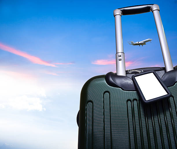 luggage and airplane on beautiful sky travel concept Bon Voyage; blank luggage tag on rolling carry-on suitcase with a beautiful blue sky backdrop and flying aircraft within the luggage pull-out handle. nice space for text. Logo or symbol can be placed on white blank tag space.  luggage tag stock pictures, royalty-free photos & images