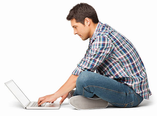 Young Man Using Laptop - Isolated Side view of a young man sitting cross-legged on floor while using laptop. Horizontal shot. Isolated on white. legs crossed at knee stock pictures, royalty-free photos & images