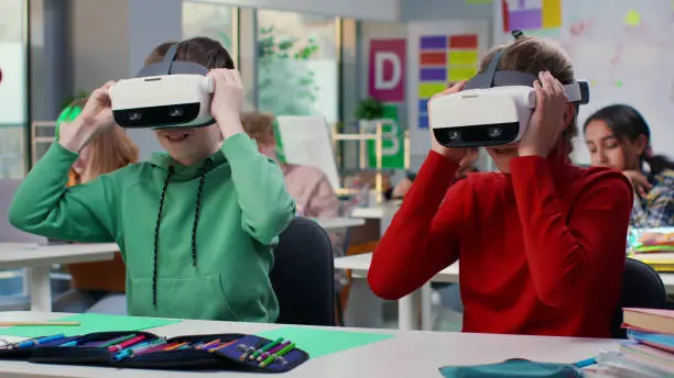 Portrait of schoolkids sitting at desk and putting on vr headset in classroom. Teenage students having modern technology lesson using virtual reality glasses