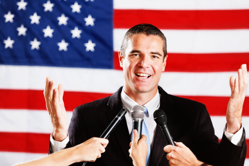 This handsome, confident-looking candidate, probably for political office, stands in front of the US flag gesturing positively and smiling, as he faces many microphones being held up eagerly by the Press.