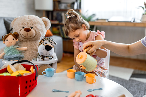 Mother and her cute little girl having a tea party with teddy and other toys at home. Family time - playing together with food blocks toys.