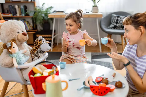 Mother and her cute little girl having a tea party with teddy and other toys at home. Family time - playing together with food blocks toys.