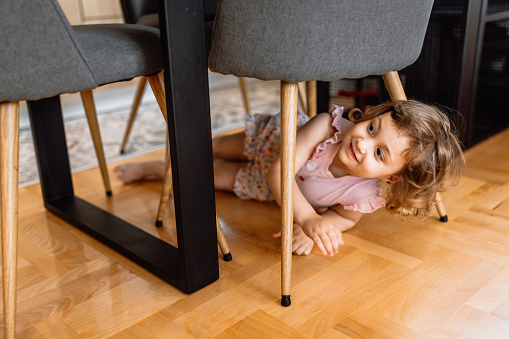 Cute little girl playing hide and seek at home, hiding under the table