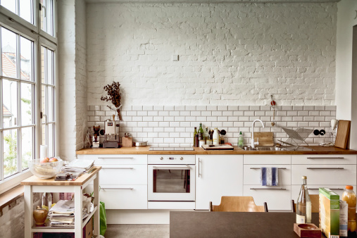 Inviting European-style kitchen illuminated by a wall of windows.  Room is comfortably cluttered with papers stacked on a side table and several bottles and packages on the table.  Fashionably distressed brick walls, painted white, give the room a rough, rustic feel.