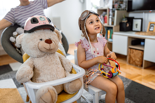 Little girl with pilot cap and her mother playing pilots at home. Little girl and her teddy bear piloting imaginary plane
