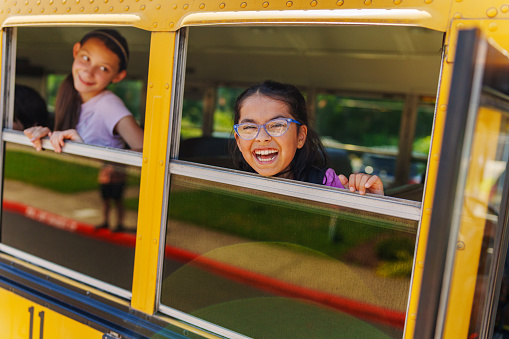 A cute elementary age Hispanic girl stands to look out the open window of a school bus and smiles directly at the camera while riding to school with friends.