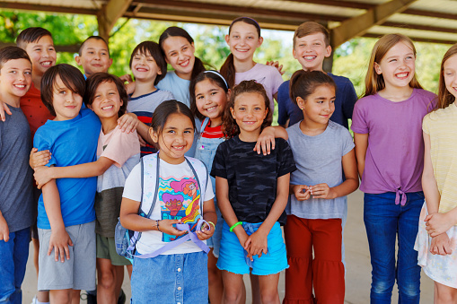 A large diverse group of elementary and middle school students huddle in closely to one another as they pose for a portrait. They are smiling as they spend time together outside in their schoolyard.