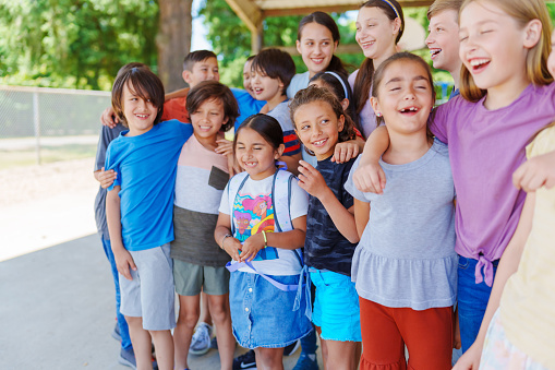 A large multiracial group of kids and teenagers huddle together with arms around each other and laugh while spending time together outside at school.