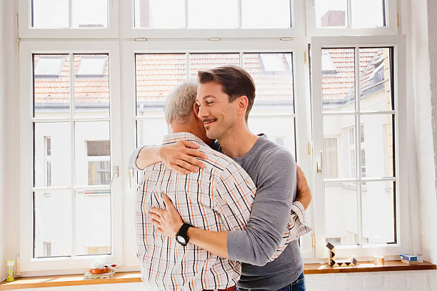 Adult son hugging his dad Adult son hugging his dad with a smile in front of the kitchen window. mercy hug stock pictures, royalty-free photos & images