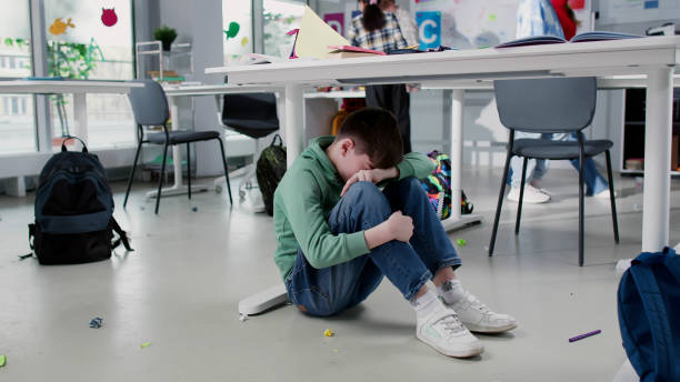 Depressed lonely boy sitting under desk in classroom with classmates playing on background