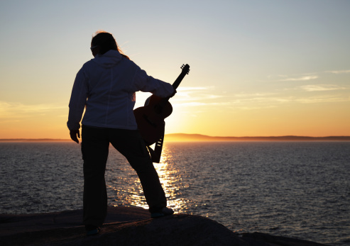 A confident woman musician back lit and silhouetted near the end of a bright sunset is holding her guitar resting on her leg.