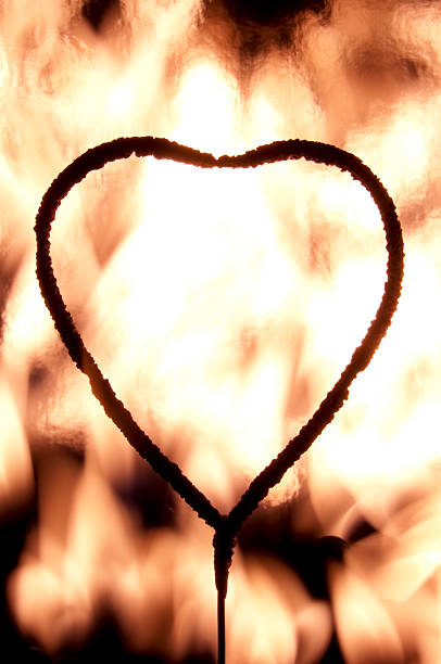 Silhouetted Heart shaped Sparkler with flames in the background.